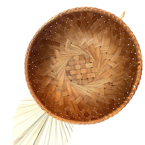 May include: A round, woven basket with a spiral design in the center. The basket is made of natural-colored, interwoven reeds. The rim of the basket is a darker shade of brown. The basket is empty and sits on a white background.