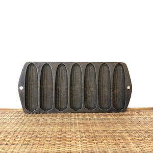 May include: A cast iron cornbread pan with seven corn cob molds. The pan is dark gray and has a rectangular shape with rounded edges and small handles. The molds are detailed to resemble corn on the cob.