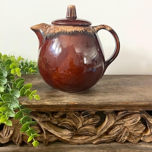 May include: A brown ceramic teapot with a lid and handle. The teapot has a glossy finish with a lighter brown band around the top. The teapot is sitting on a wooden surface.