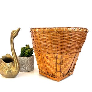 May include: A woven basket with a square base and a round top. The basket is made of light brown wicker and has a natural, rustic appearance. A brass swan-shaped container and a small potted plant are to the left of the basket.