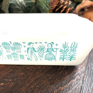 May include: Off-white rectangular casserole dish with a turquoise illustration of people, plants, and baskets. The dish has a slightly curved edge and is set on a dark wooden surface. The design is reminiscent of vintage kitchenware.