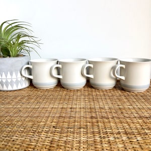 May include: Set of four white ceramic mugs with a light blue band around the top. The mugs are arranged in a row on a woven wicker surface.