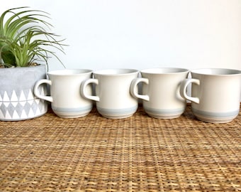 May include: Set of four white ceramic mugs with a light blue band around the top. The mugs are arranged in a row on a woven wicker surface.