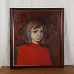 May include: A framed portrait of a young person with shoulder-length brown hair and a red top. The painting has a dark red background and is set in a dark brown frame with a decorative border. The artwork is displayed on a wooden surface.