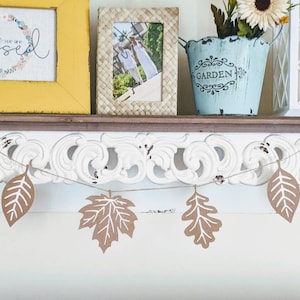 May include: A decorative shelf with a yellow framed picture that says "blessed", a woven framed photo, a blue bucket with flowers, and a white lantern. A garland of brown paper leaves hangs below.