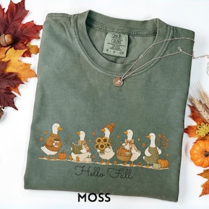May include: An olive green t-shirt featuring a graphic of five white ducks engaged in autumn activities, including holding a coffee cup, a book, sunflowers, a basket, and wearing an apron. The text "Hello Fall" is printed below the ducks.
