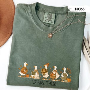 May include: A moss green t-shirt featuring a graphic of five white ducks dressed in autumn attire, including sweaters, scarves, and hats, with pumpkins and sunflowers. The text "Hello Fall" is printed in black script below the ducks.