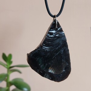 Op de afbeelding: Een zwarte obsidiaanhanger met een glad, gepolijst oppervlak. De hanger heeft een onregelmatige vorm en hangt aan een zwart koord. De hanger is ongeveer 5 cm lang.