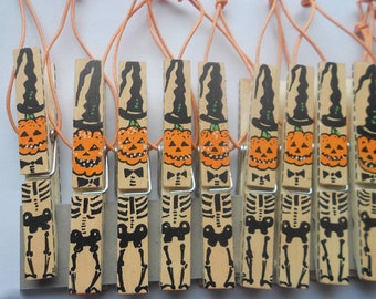 Könnte beinhalten: Eine Sammlung von Holz-Wäscheklammern zum Thema Halloween. Jede Klammer zeigt ein handgemaltes Design eines Kürbisses mit Hexenhut und eines Skeletts. Die Klammern sind durch eine orangefarbene Schnur verbunden.