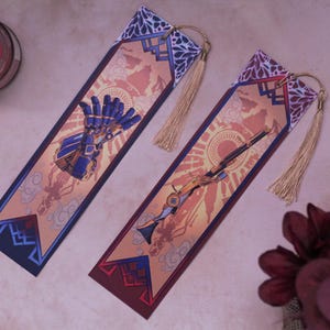 Può includere: Due segnalibri decorativi con nappe dorate. Uno presenta una mano robotica blu e oro, l'altro un'arma. Entrambi hanno una combinazione di colori rosso, arancione e blu con un disegno di sole e nuvole.