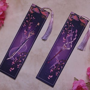 May include: Two purple and white bookmarks with tassel ends. Each bookmark features a different illustration of a weapon, with cherry blossom branches and cloud designs. The bookmarks are rectangular and have a dark purple background.
