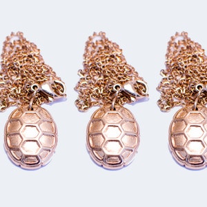 Puede incluir: Tres collares de tono dorado con un colgante en forma de tortuga. Cada colgante tiene un patrón hexagonal texturizado.
