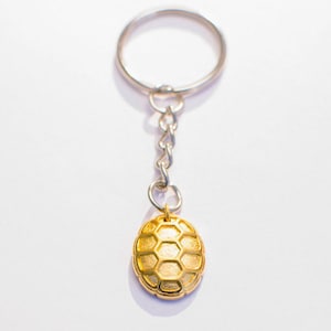Op de afbeelding: Een gouden sleutelhanger met een voetbalontwerp. De sleutelhanger is bevestigd aan een zilveren metalen ring en ketting.