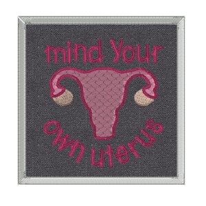 May include: Embroidered patch with a pink uterus and the text "mind your own uterus" in pink thread on a dark gray background.