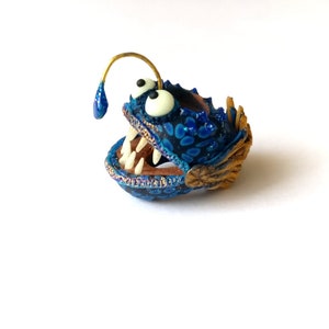 Könnte beinhalten: Eine blaue und goldene Anglerfisch-Figur mit einem großen Maul und scharfen Zähnen. Die Figur hat einen kleinen, goldenen Köder auf dem Kopf.