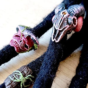 Puede incluir: Primer plano de rastas negras adornadas con abalorios decorativos de calaveras y arañas. Las calaveras están pintadas en tonos rojos, rosas y morados, mientras que la araña es verde y marrón. Los abalorios añaden un toque gótico único al peinado.