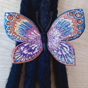 Puede incluir: Una colorida pinza para el pelo con forma de mariposa, con alas naranjas, azules y moradas. La mariposa tiene un acabado brillante y está sujeta a una base negra. El accesorio para el pelo está diseñado para rastas.