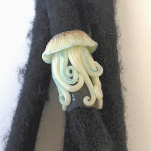 Op de afbeelding: Een lichtblauwe en witte kwallen-vormige kraal met een zwarte basis. De kraal is bevestigd aan een zwarte dreadlock.