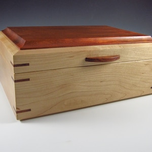 May include: A wooden box with a hinged lid and a small wooden handle. The box is made of maple wood with a mahogany top. The box has a simple design and is perfect for storing small items.
