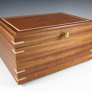 May include: A wooden box with a hinged lid and a brass knob. The box is made of a light brown wood with a darker brown wood inlay around the edges. The box is sitting on a white surface.