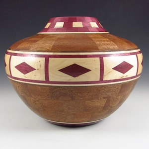 May include: A wooden vase with a geometric pattern of red, white, and brown wood. The vase is round and has a wide opening.