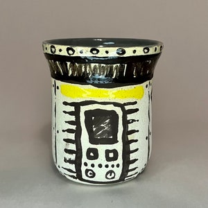 May include: A white ceramic mug with black and yellow geometric designs. The mug has a black rim and a black band around the top. The design features a stylized window with a black frame and a black rectangle inside.