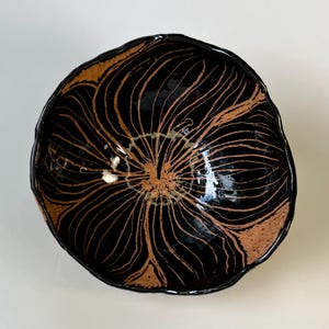 May include: A handmade ceramic bowl with a unique design. The bowl is primarily black with a glossy finish and features a brown, floral-like pattern radiating from the center. The rim has a slightly irregular shape.