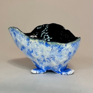 May include: A blue and white ceramic bowl with a black glaze on the inside. The bowl has a unique, organic shape and three legs.