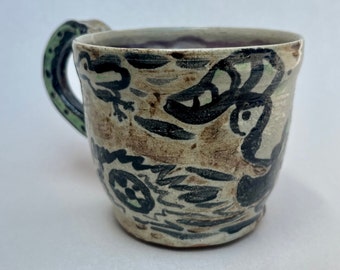 May include: A ceramic mug with a green handle and a white glaze. The mug is decorated with black and brown abstract designs.