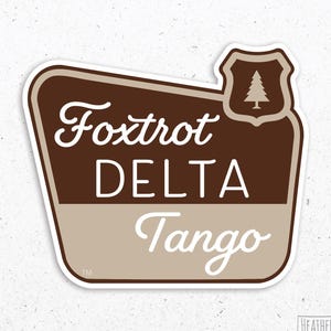 May include: A brown and beige sticker with a white outline. The sticker features a stylized design of a road sign with the text "Fox Trot Delta Tango" and a small pine tree icon in the upper right corner.