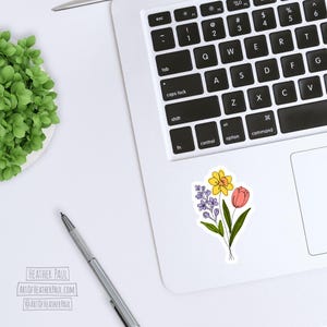 sticker featuring a purple freesia, a yellow daffodil with and orange center and a  pinkish-red tulip mocked up on a laptop computer