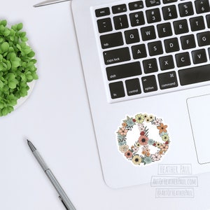 May include: A peace sign sticker with a floral design. The peace sign is surrounded by flowers in shades of pink, yellow, blue, and green. The sticker is on a white laptop.