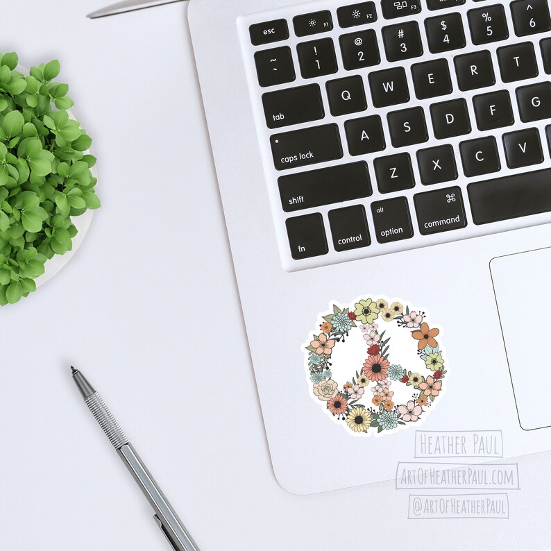 May include: A peace sign sticker with a floral design. The peace sign is surrounded by flowers in shades of pink, yellow, blue, and green. The sticker is on a white laptop.