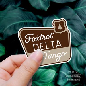 sticker featuring a national parks style sign with the text reading the phrase foxtrot delta tango over a background with green leaves