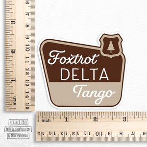 sticker featuring a national parks style sign with the text reading the phrase foxtrot delta tango over a white background with brown rulers to show sizing