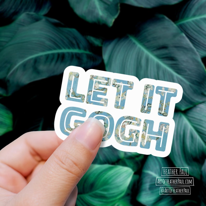May include: A white sticker with the text "Let it Gogh" in blue letters. The letters are filled with a blue and white floral pattern. The sticker is being held in a hand against a green leafy background.