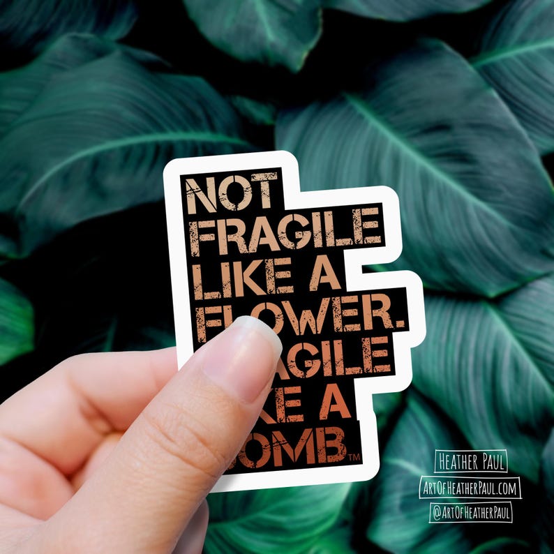 sticker reading "not fragile like a flower. fragile like a bomb" in a warm gradient over a background with green leaves
