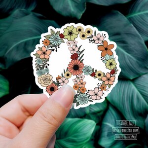 May include: A white peace sign sticker with a floral design. The flowers are in shades of pink, yellow, orange, and green. The sticker is on a green leafy background.