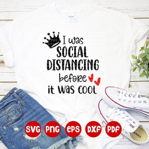 May include: White t-shirt with black text that reads "I was social distancing before it was cool" with a crown above the text and two red hearts below the text. The t-shirt is on a wooden surface with a pair of white sneakers and blue denim shorts.