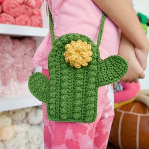 Puede incluir: Un bolso de mano de color verde tejido a crochet con forma de cactus y un detalle de flor amarilla. El bolso lo lleva puesto un niño.