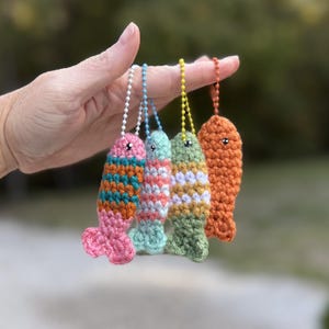 May include: Four small, crocheted fish keychains hanging from individual ball chains. The fish are multicolored with stripes in shades of pink, teal, orange, light blue, white, and green, each featuring a single small black bead for an eye.