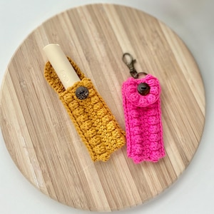 Pode incluir: Dois estojos de palha de crochê, um amarelo mostarda e outro rosa chiclete, com botões de madeira. Os estojos estão sobre uma tábua de cortar de madeira.