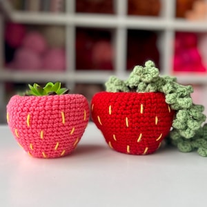 Puede incluir: Dos macetas de fresa de ganchillo, una rosa y otra roja, con detalles de costura amarillos. La maceta rosa contiene una pequeña suculenta verde. Una planta verde en forma de vid sale de la maceta roja.