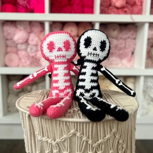 Op de afbeelding: Twee gehaakte skeletpoppen, een roze en een zwarte, zitten op een gevlochten kruk. De poppen hebben witte ogen en een witte ribbenkast.