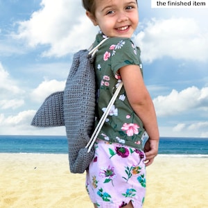 May include: A young girl wearing a green and floral shirt and pink floral shorts is wearing a grey crocheted shark backpack. The backpack has a white drawstring. The girl is standing on a sandy beach with the ocean in the background. Listing is for the PDF instant digital download of the shark backpack crochet pattern modification. Not the finished item.