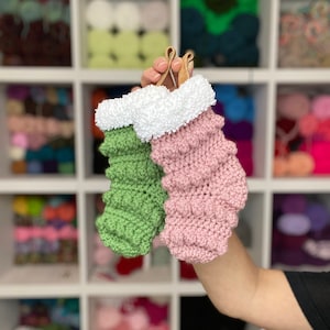 May include: Two small crocheted Christmas stockings, one green and one pink, with white faux fur trim. The stockings are held in a hand.