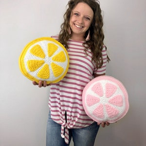 May include: Two crocheted lemon slice pillows, one yellow and one pink, held by a person wearing a pink and white striped shirt and blue jeans.