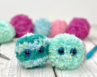 May include: Two fuzzy, green and blue yarn balls with googly eyes. The yarn balls are sitting on a white wooden surface with pink and blue yarn balls in the background.