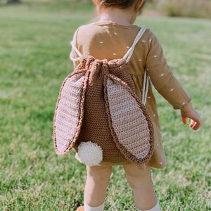 May include: A young child wearing a brown crocheted bunny backpack with a white pom-pom tail. The child is standing in a grassy field with a blue sky in the background.