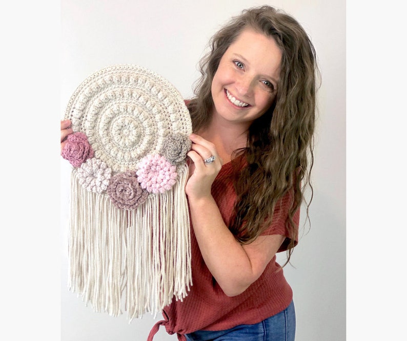 Puede incluir: Un tapiz de pared de crochet blanco con un dise&ntilde;o circular y flores de crochet rosas y grises. El tapiz de pared tiene una larga franja blanca.